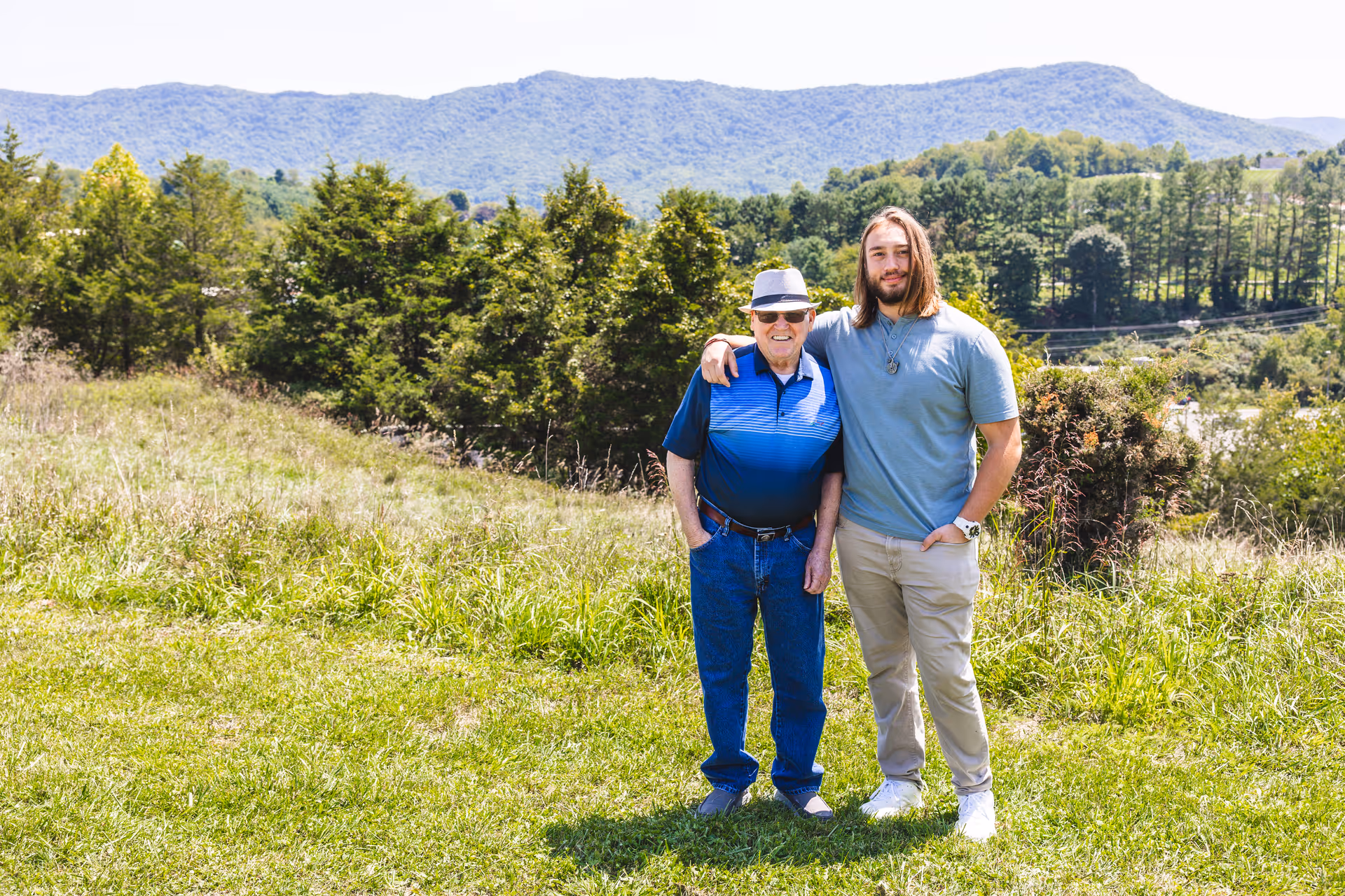 An elderly man wearing a hat and sunglasses stands outdoors on a grassy area with a younger man who has long hair. The younger man has his arm around the elderly man's shoulder. Behind them are trees and hills under a clear sky.