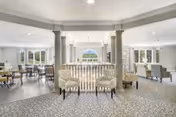 A spacious and well-lit common area in a senior living facility featuring multiple seating arrangements with chairs and tables. The room has large windows allowing natural light to fill the space, decorative columns, and a patterned carpet. The overall ambiance is bright and welcoming.
