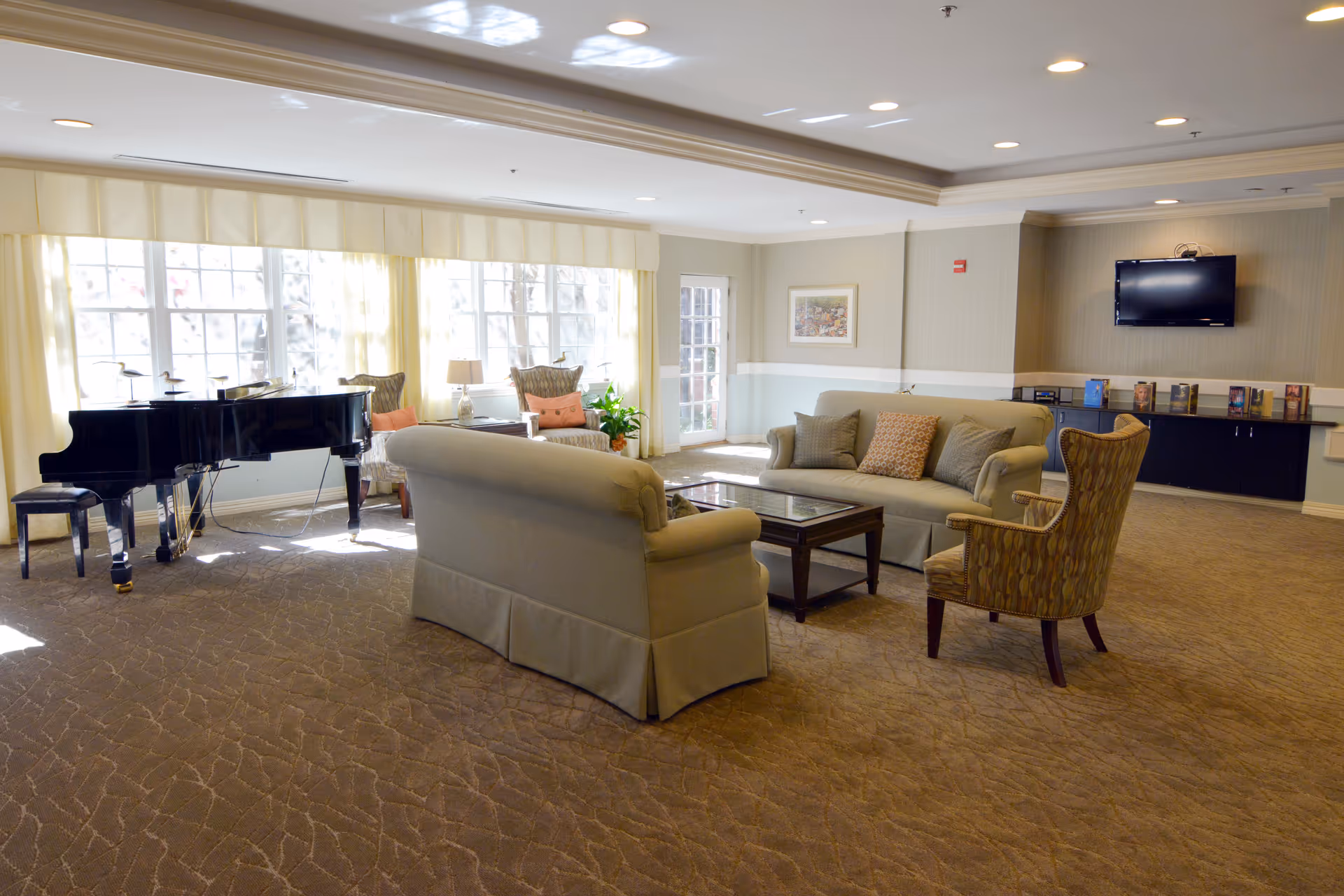 Bright lounge with sofas, armchairs, a grand piano by large windows, and a wall-mounted TV.