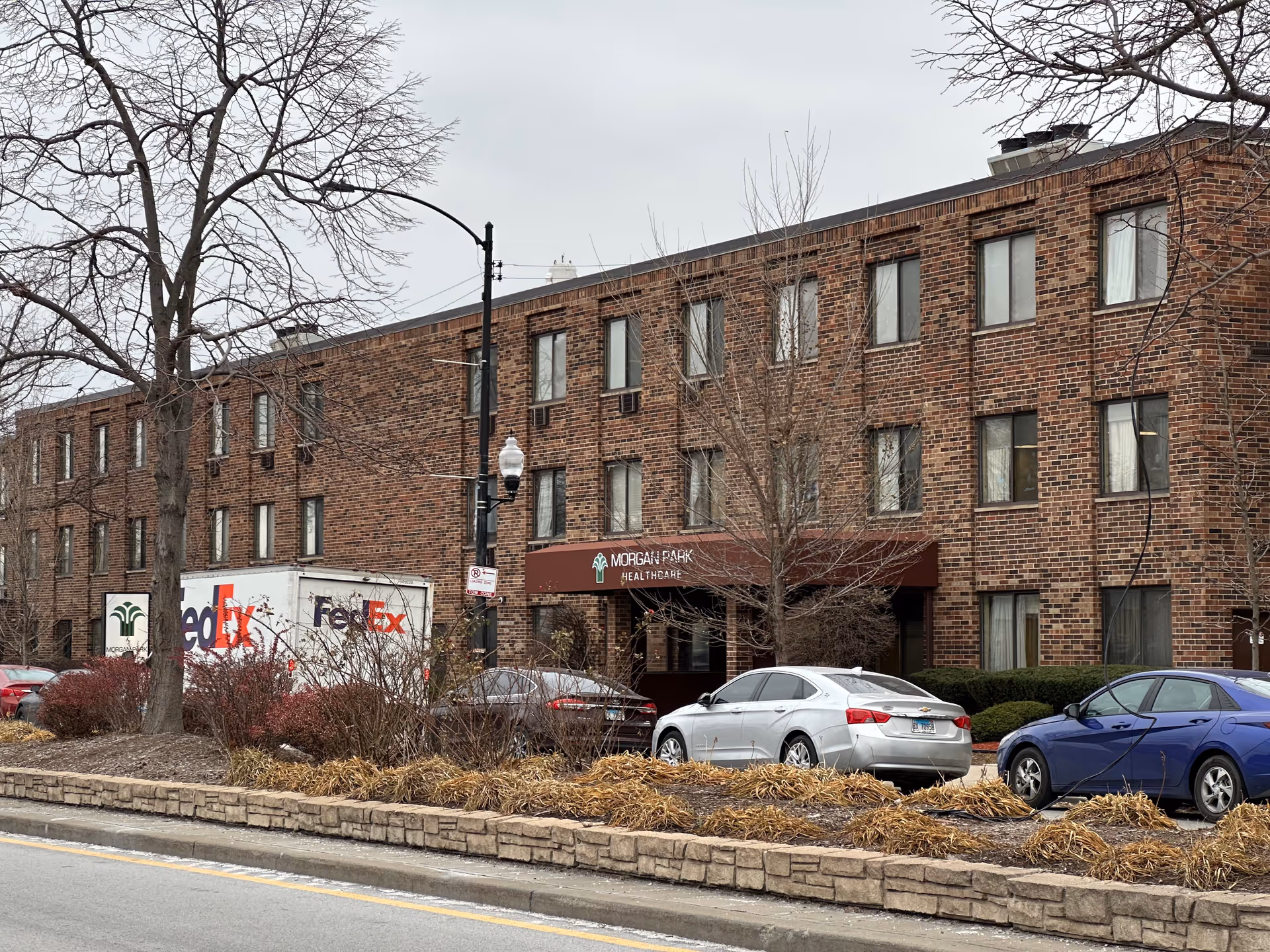 Exterior view of a three-story brick building with multiple windows, a brown awning labeled 'Morgan Park Healthcare', several parked cars, a FedEx truck, leafless trees, and a stone-lined planting bed along the street.