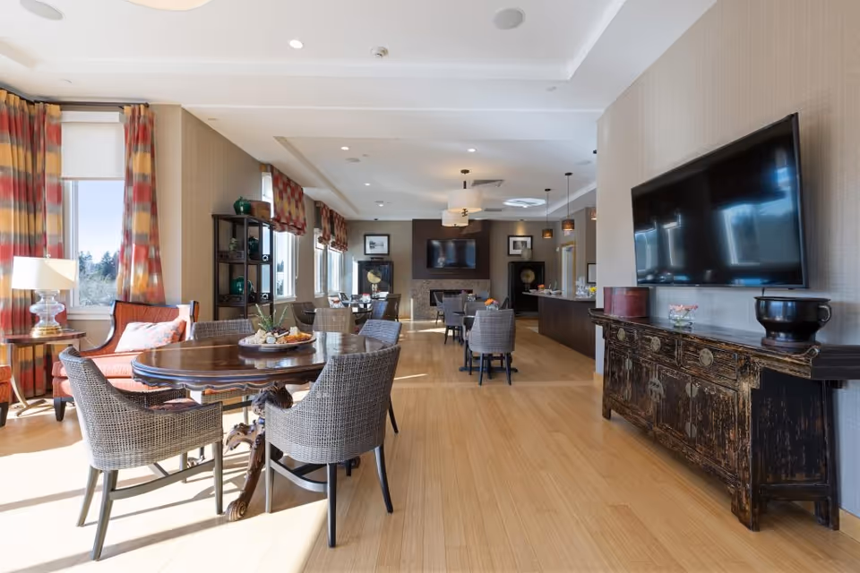 Sunlit communal dining and lounge area with round wooden tables and wicker chairs, a large wall-mounted TV, and windows with colorful curtains.
