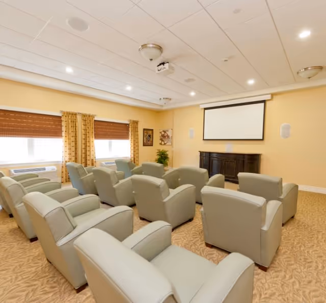 A cozy media room with multiple beige recliner chairs arranged in rows facing a large projector screen mounted on a light yellow wall. The room has carpeted flooring, two windows with brown blinds and patterned curtains, and ceiling lights.