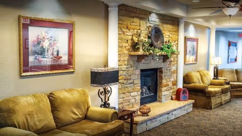 A cozy living room area with two mustard-colored sofas, a stone fireplace decorated with plants and a decorative plate, a side table with a lamp, framed floral artwork on the walls, and a ceiling fan with lights.