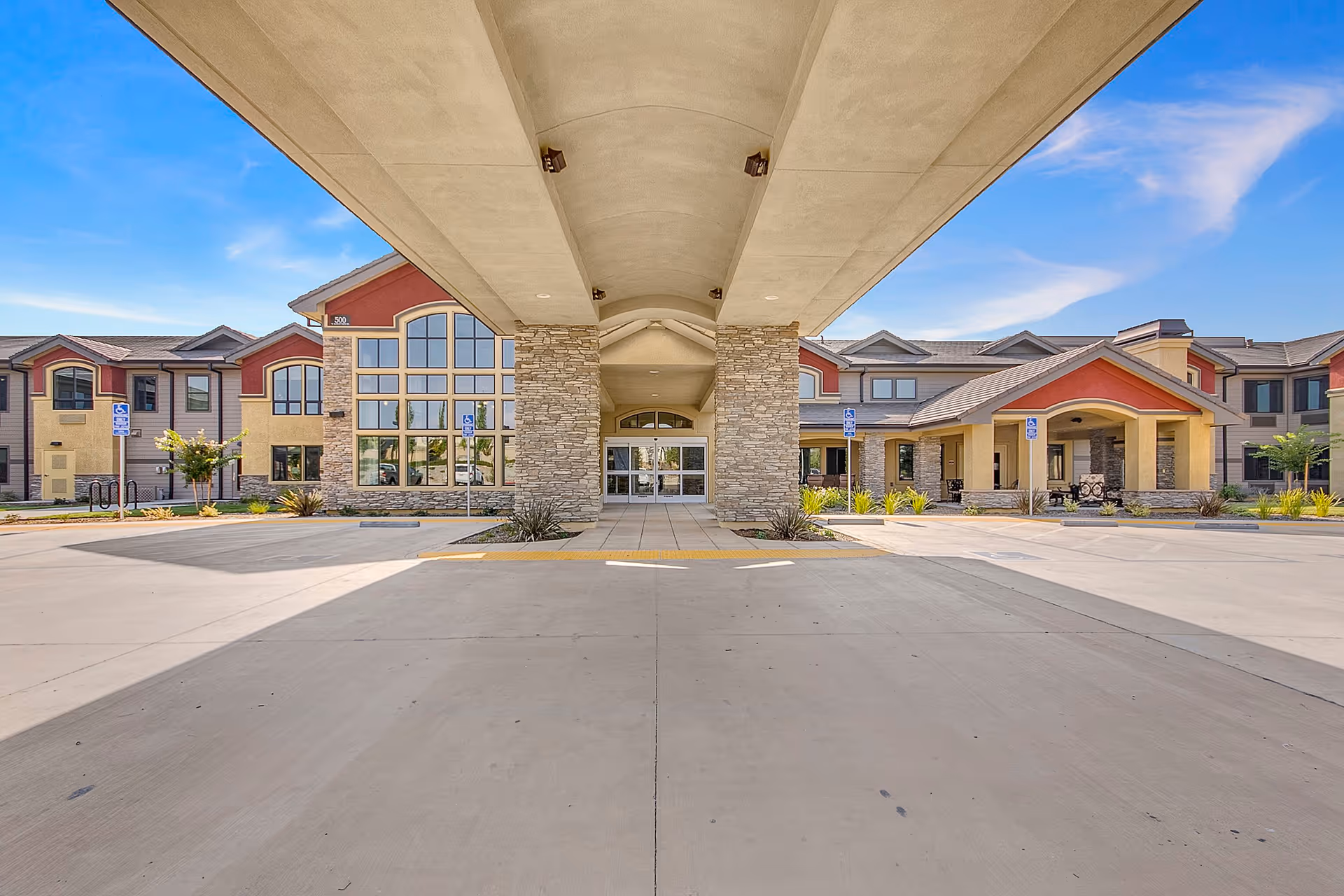 Front exterior view of The Pines, A Merrill Gardens Community building with a covered entrance, stone pillars, large windows, and a clear blue sky.