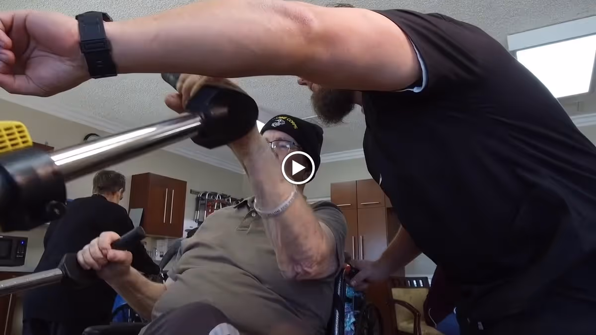 An elderly man wearing a beanie and glasses is exercising with a resistance machine while a caregiver assists him in a room with kitchen cabinets in the background. Another person is visible in the background near the kitchen area.