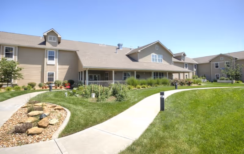 Exterior view of a senior living facility building with beige siding and a sloped roof under a clear blue sky. There is a well-maintained lawn with a curved concrete walkway, landscaped garden beds with rocks and plants, and small outdoor lights along the path.