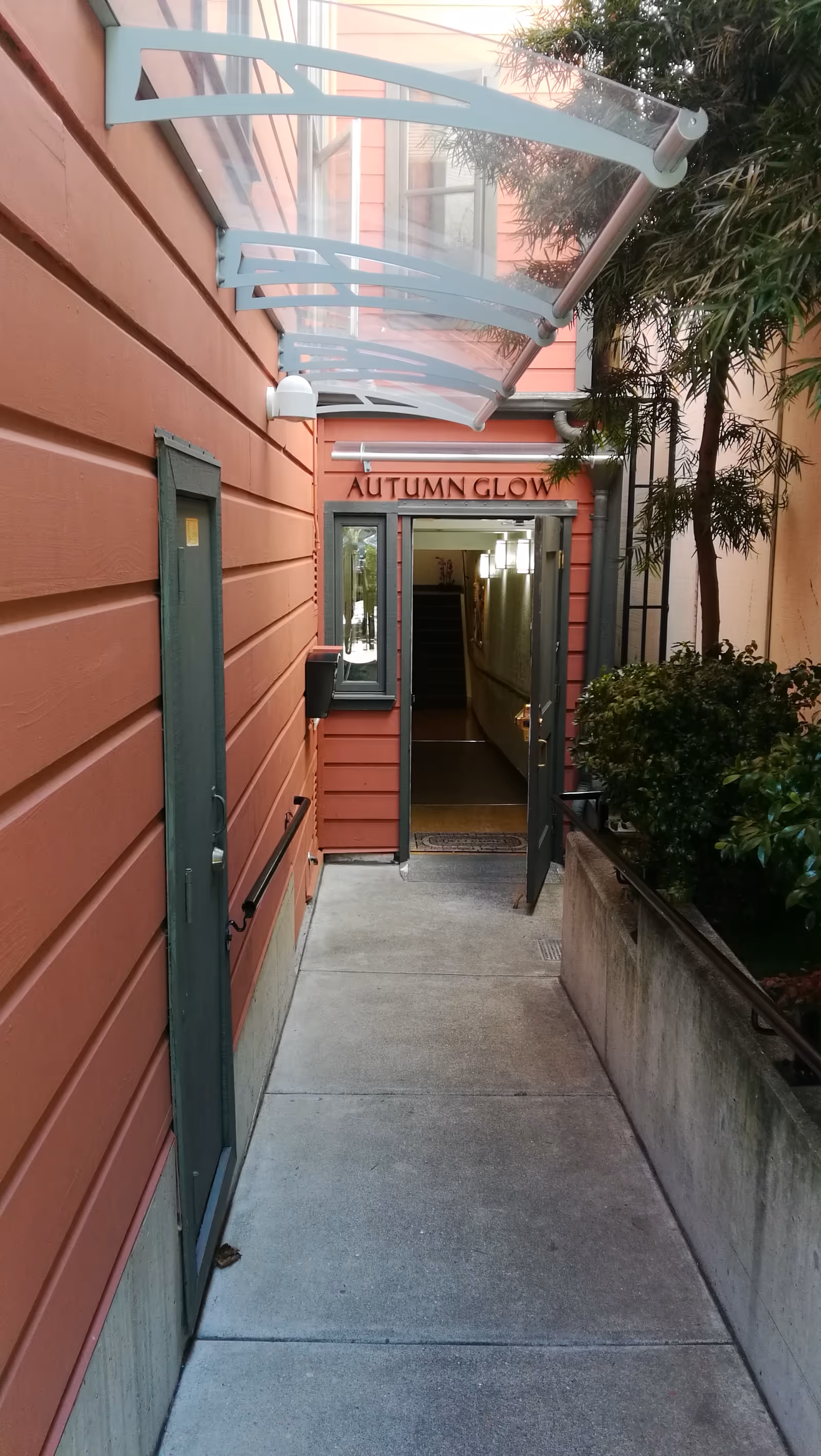 Narrow outdoor walkway leading to an entrance door with the sign 'AUTUMN GLOW' above it. The building exterior is painted in a reddish color with green trim around the door and window. There is a transparent awning above the walkway and some greenery on the right side.