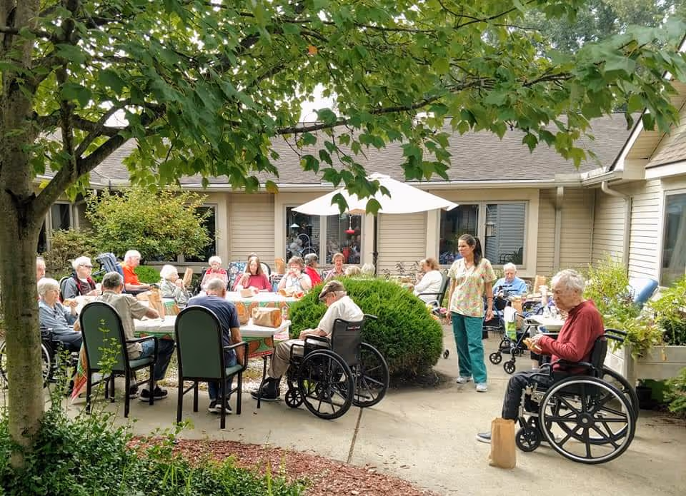 A group of elderly people sitting outdoors around tables under a large white umbrella in a courtyard area of a senior living facility. Some individuals are in wheelchairs, and a caregiver is standing nearby. The area is surrounded by greenery and beige buildings.