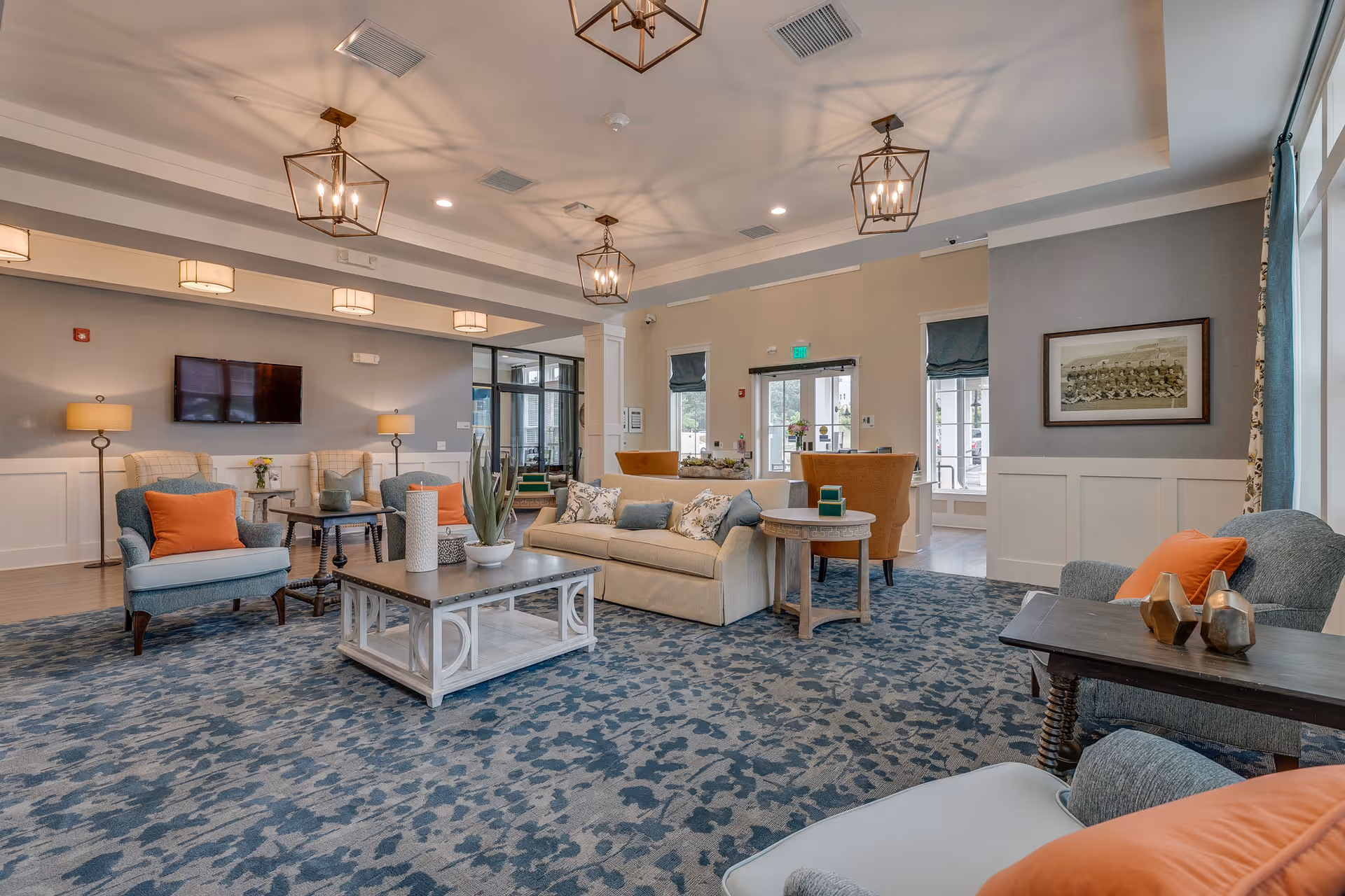 A spacious and well-lit living room area in Everlan of Clemson featuring multiple seating options including armchairs with orange cushions, a beige sofa with patterned pillows, and side tables. The room has a patterned blue carpet, modern pendant lights hanging from a tray ceiling, a wall-mounted TV, and large windows with teal curtains allowing natural light to enter.