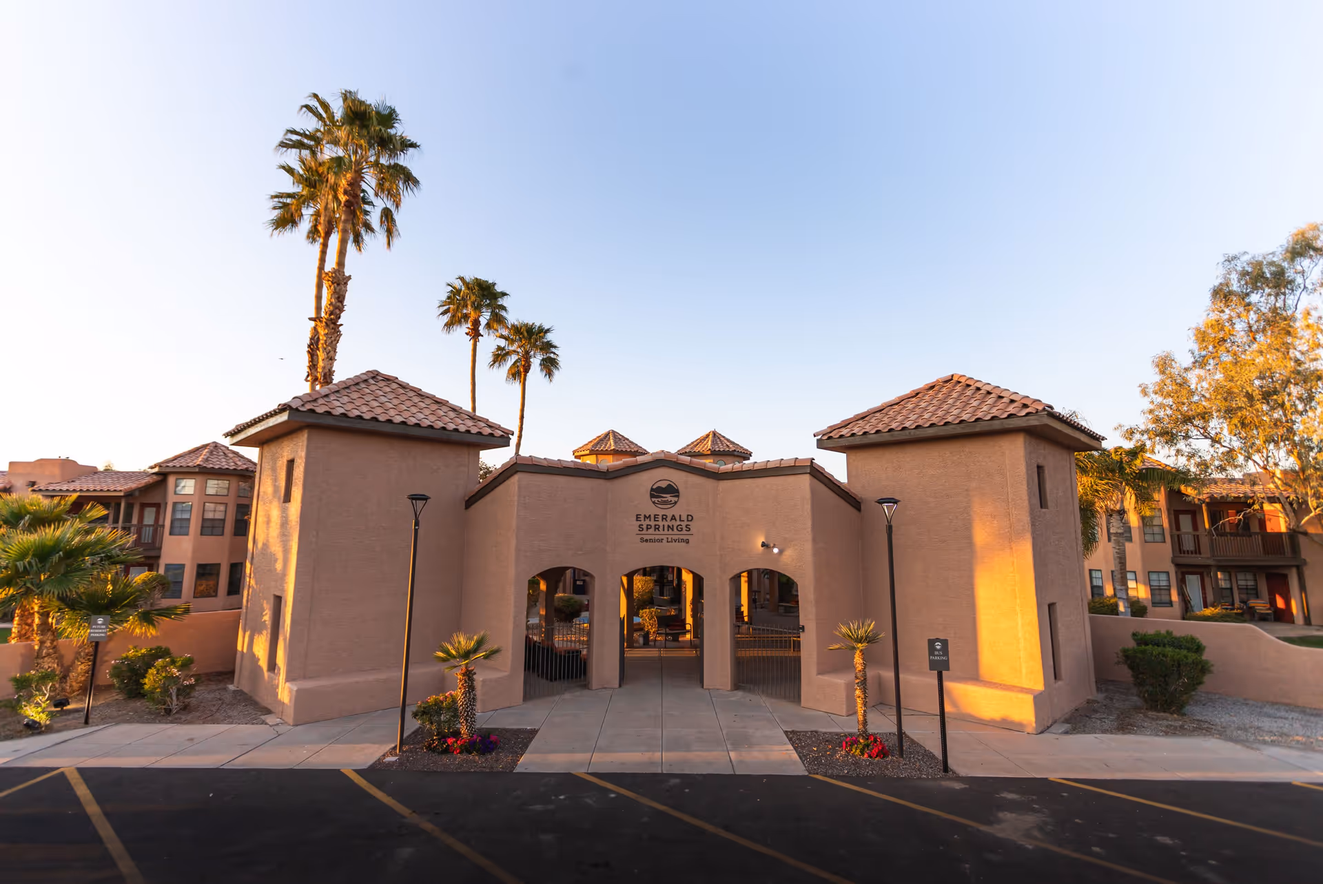 Exterior view of the entrance to Emerald Springs Senior Living facility with a beige stucco building featuring three archways, tiled roofs, palm trees, and landscaped plants under a clear blue sky.