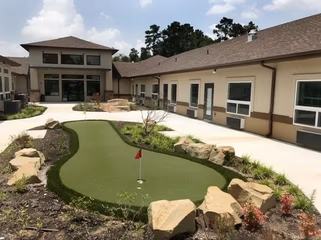 Outdoor courtyard area at Crimson Heights Health & Wellness featuring a small putting green surrounded by rocks and landscaping, with a bench and building entrances visible in the background under a partly cloudy sky.
