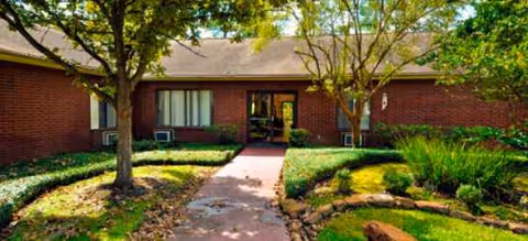 Brick senior living building exterior with a central entrance, walkway, and landscaped greenery.