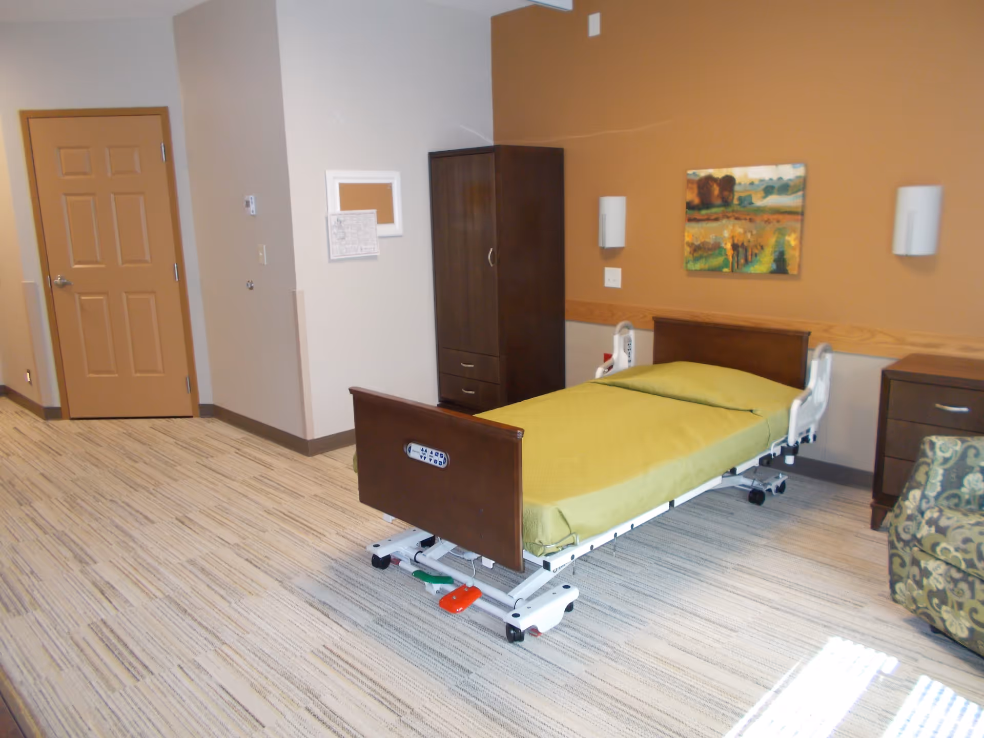 A senior living facility bedroom with a hospital-style bed covered with a green blanket. The room has beige walls, a wooden wardrobe, a nightstand, a patterned armchair, and a landscape painting on the wall. The floor is carpeted with a striped pattern, and there is a closed wooden door in the background.