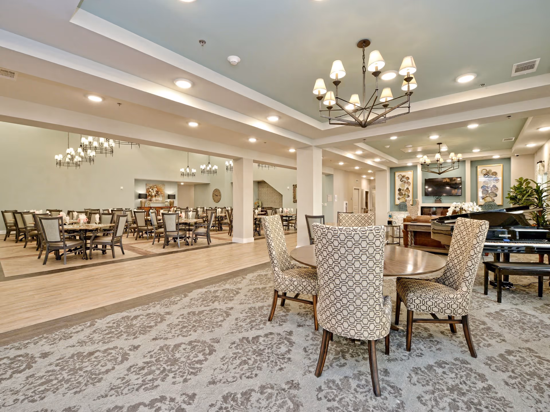 A spacious and well-lit dining and lounge area in a senior living facility. The room features multiple dining tables with chairs, a patterned carpet, a grand piano, and a wall-mounted TV. The walls are decorated with framed art and plants, and the ceiling has recessed lighting and chandeliers.