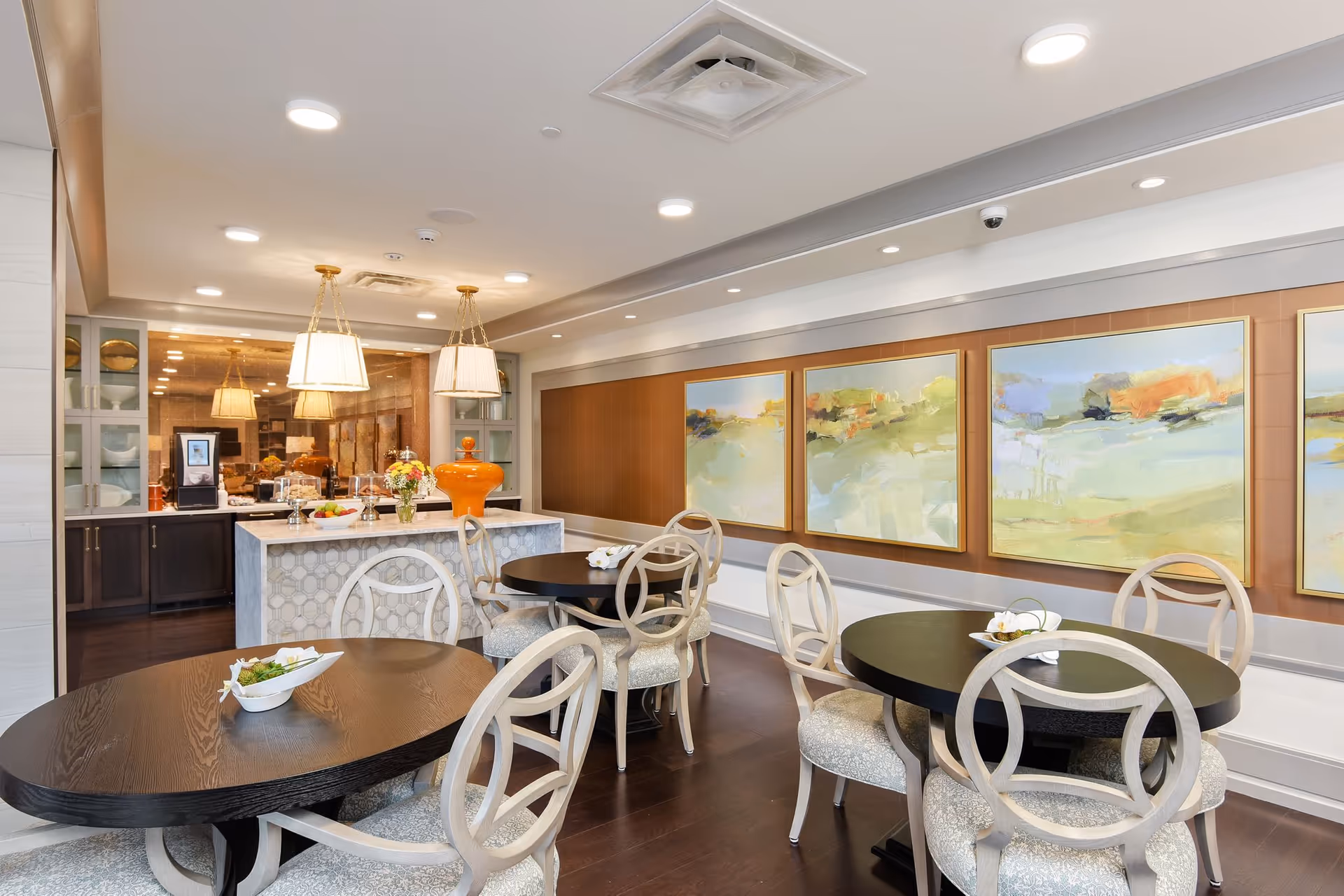 A bright and modern dining area with round black tables and white cushioned chairs. The room features a small kitchen or serving area with a marble island topped with decorative items and a coffee machine. The walls are adorned with large abstract landscape paintings in soft colors. The ceiling has recessed lighting and two hanging pendant lamps above the island.