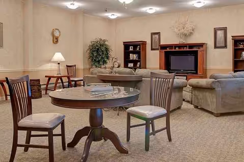 A cozy senior living lounge with round table, chairs, sofas, and a TV.