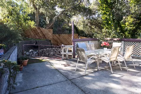Sunlit concrete patio with a glass-top table, metal chairs, potted plants, and lattice fencing backed by trees.