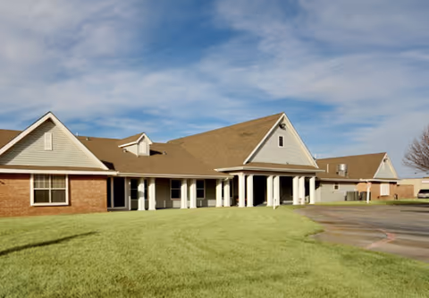 Exterior view of a single-story building with a combination of brick and siding walls, a large covered entrance supported by white columns, and a well-maintained lawn under a partly cloudy sky.
