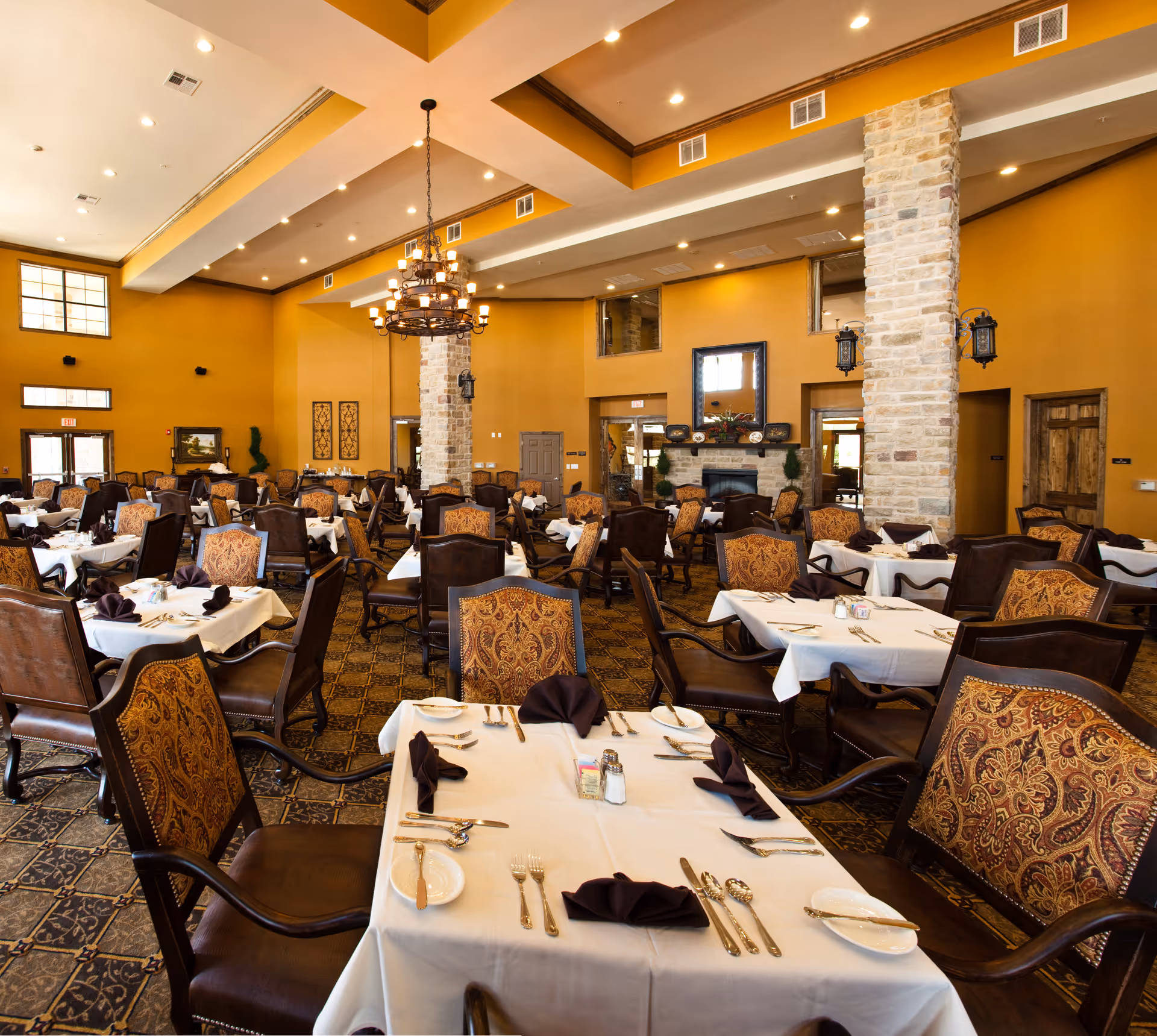 Spacious dining room with multiple tables covered in white tablecloths, set with silverware, plates, and folded dark napkins. The room features high ceilings with recessed lighting, a large chandelier, stone pillars, and warm yellow walls. Upholstered chairs with ornate patterns surround the tables, and a fireplace with a mirror above it is visible in the background.