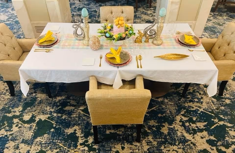 A beautifully set dining table with a white tablecloth, featuring decorative plates, utensils, and a centerpiece of flowers and fruits.