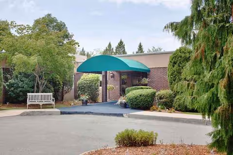 Senior living facility entrance with brick exterior, bench, teal awning, and surrounding greenery.