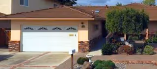 Exterior view of a single-story house with a white garage door, a tiled roof, and a small front yard featuring bushes and a tree.