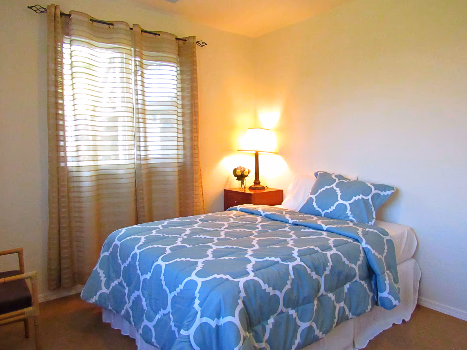 A cozy bedroom with a bed covered in a blue and white patterned comforter and matching pillow. A wooden nightstand with a lit lamp and a small flower vase is beside the bed. Beige curtains cover a window on the left side, and a wooden chair is partially visible in the corner.