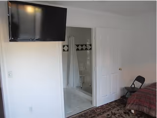 A room with a wall-mounted flat-screen TV, a partially open door revealing a bathroom with a shower curtain and a shower chair, a bed with a red and brown striped blanket, a folding chair, and a patterned rug on the floor.