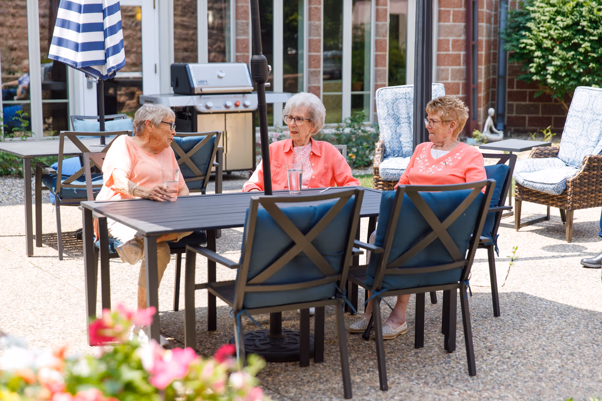 Three elderly women sitting and talking around a black outdoor table with blue cushioned chairs on a patio. There is a striped umbrella, a grill, and wicker chairs with blue and white cushions in the background. The setting is sunny with some greenery and flowers visible.
