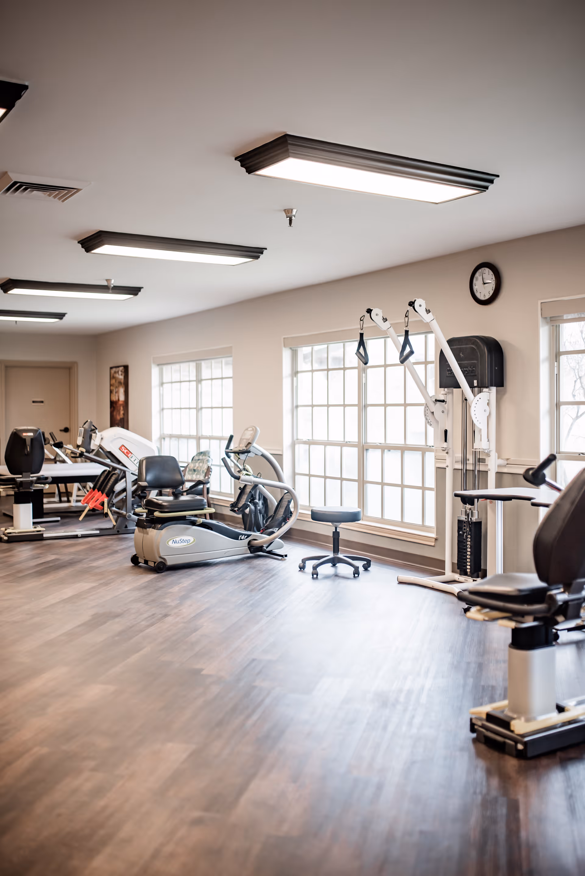 A bright exercise room with large windows letting in natural light. The room contains various pieces of fitness equipment including recumbent bikes, a multi-function pulley machine, and a rolling stool. The floor is wooden, and the ceiling has rectangular fluorescent lights. A clock is mounted on the wall above the pulley machine.