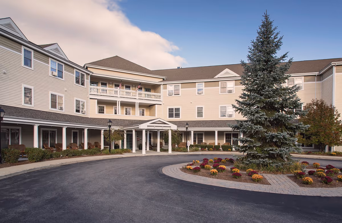 Exterior view of Traditions of Dedham senior living facility showing a three-story building with beige siding, multiple windows, a covered entrance with white columns, a circular driveway, and landscaped area with a large evergreen tree and colorful flowers.