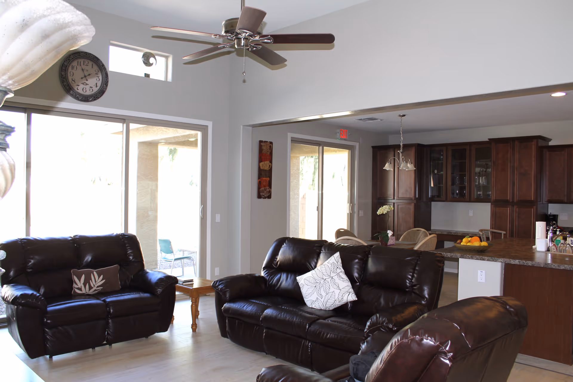 A spacious living room with dark brown leather sofas and a matching armchair. The room has light-colored walls and flooring, a ceiling fan with lights, and a large clock on the wall. Sliding glass doors lead to an outdoor patio area with chairs. In the background, there is a kitchen area with wooden cabinets, a countertop with fruit, and a dining table with chairs.