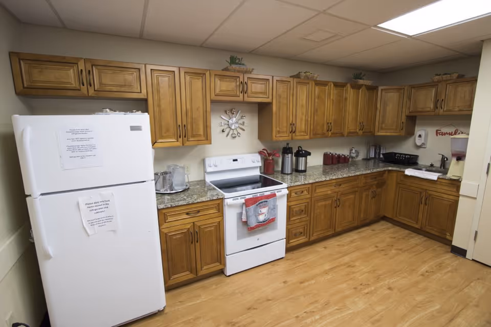 A kitchen area with wooden cabinets, a white refrigerator with notices taped on it, a white stove with a red and gray towel hanging on the handle, and a granite countertop holding various items including coffee dispensers, a utensil holder, and a soap dispenser. The floor is wooden and the ceiling has white tiles.