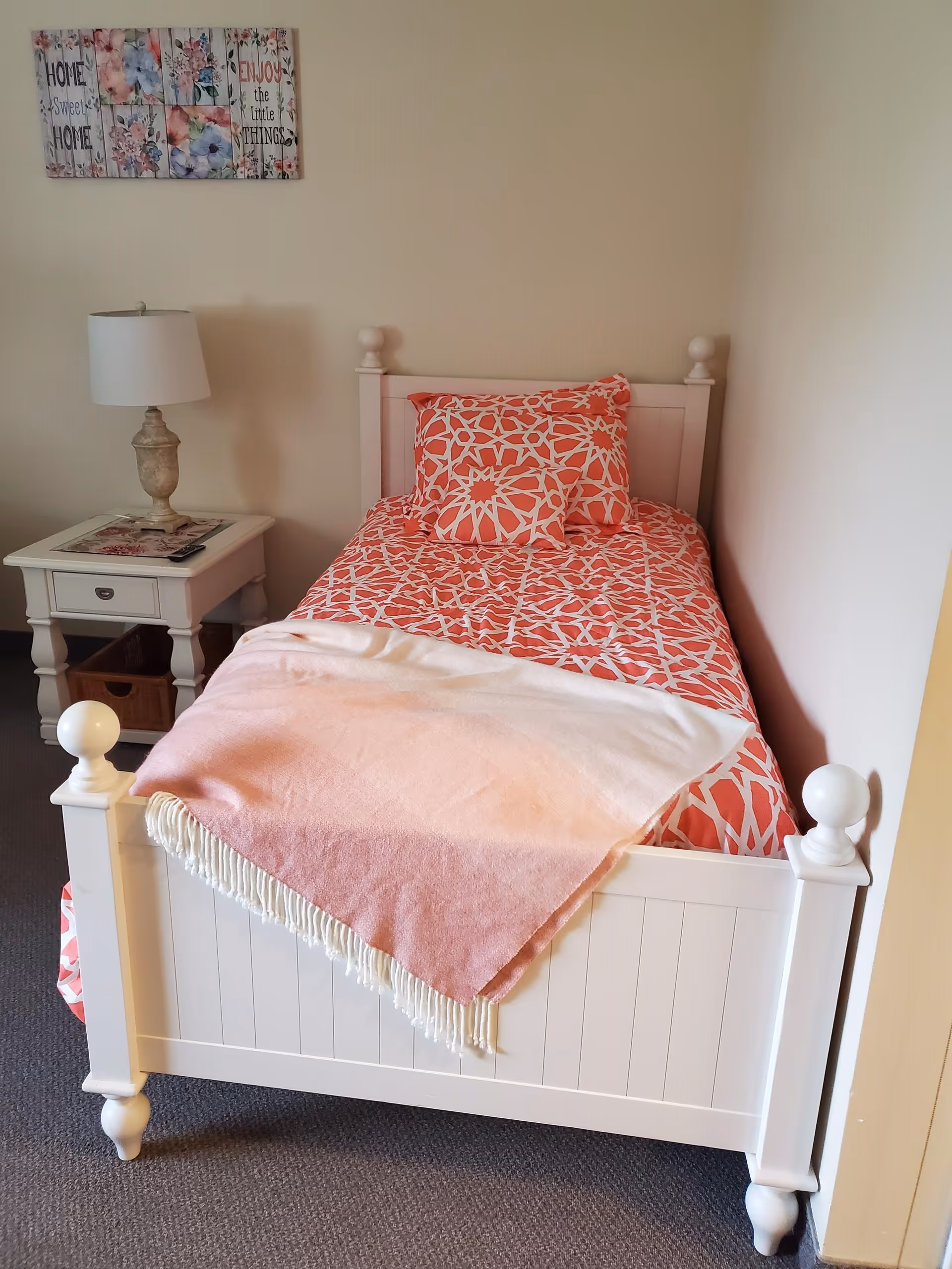 A small bedroom with a white wooden single bed featuring a coral and white patterned bedspread and matching pillows. A pink and white fringed throw blanket is draped over the foot of the bed. Next to the bed is a white nightstand with a decorative lamp and a remote control on top. Above the bed, there is a wall decoration with floral designs and the phrases 'Home Sweet Home' and 'Enjoy the Little Things'.