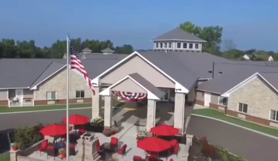 Front entrance of a single-story retirement community building with an American flag, a covered porte-cochere, and outdoor seating with red umbrellas.