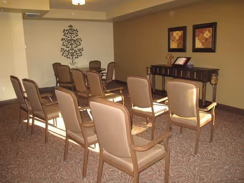 A small meeting or chapel room with two rows of beige cushioned chairs facing a wooden table with a cross, an open book, and two framed floral pictures on the wall behind it. The room has beige walls and patterned carpet flooring.