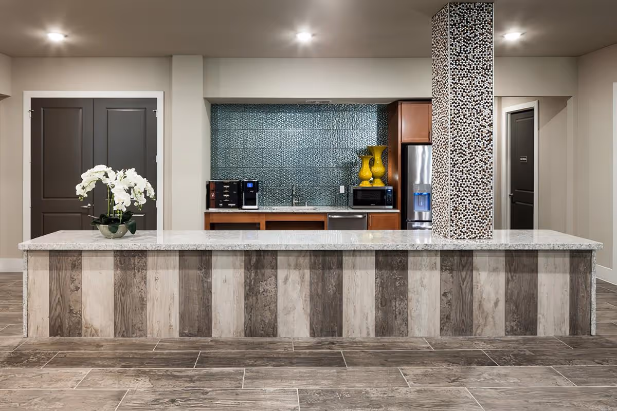 Modern kitchen area with a long marble countertop island featuring a vase with white flowers. The back wall has a blue textured backsplash, wooden cabinets, a stainless steel refrigerator, microwave, coffee machine, and two yellow decorative vases. A column covered with mosaic tiles is integrated into the countertop. The floor has wood-like tiles and there are two dark-colored doors on either side of the kitchen area.
