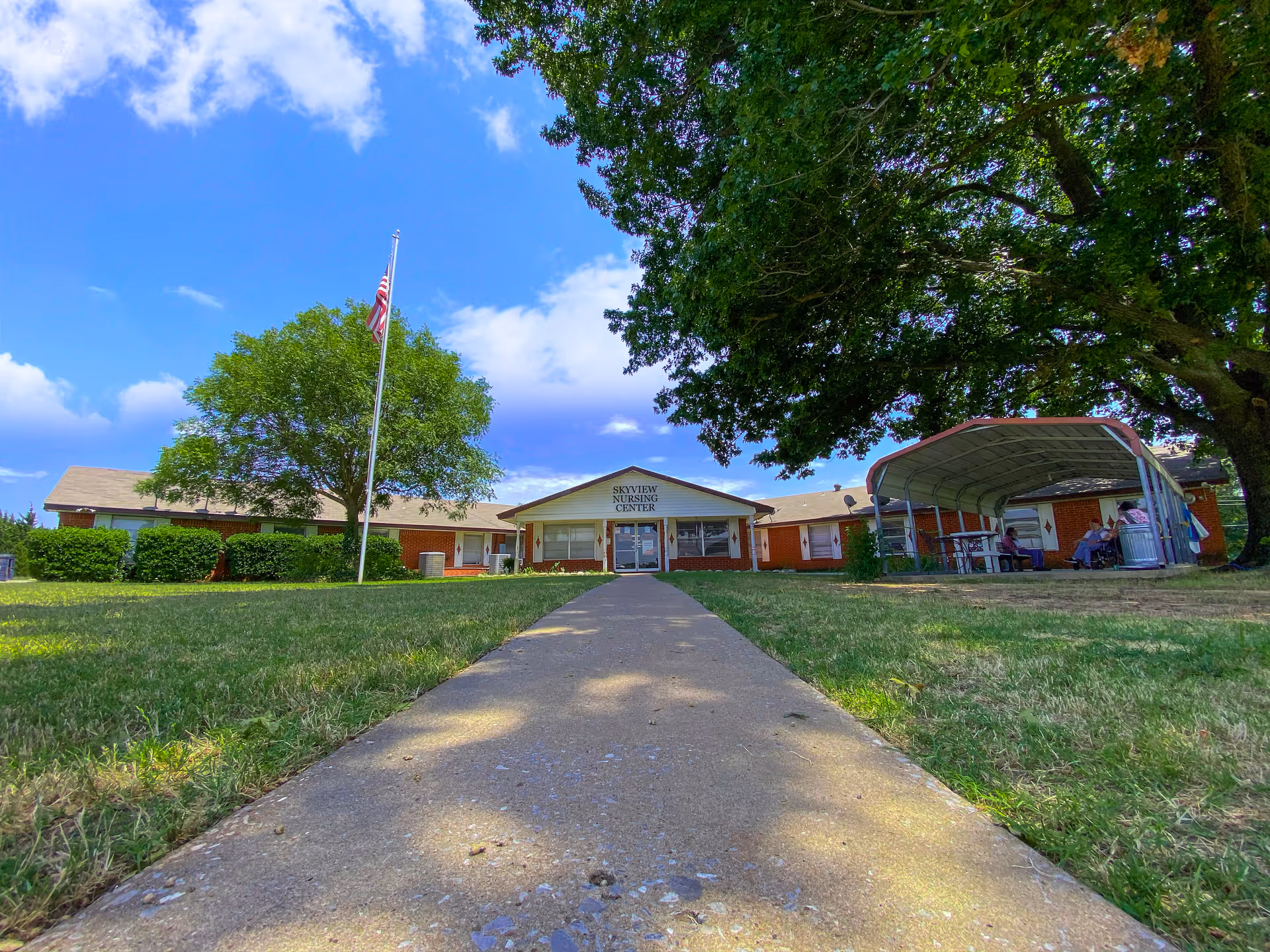 Front exterior of Skyview Nursing Center with a central walkway, flagpole, large trees, and a covered outdoor seating area.