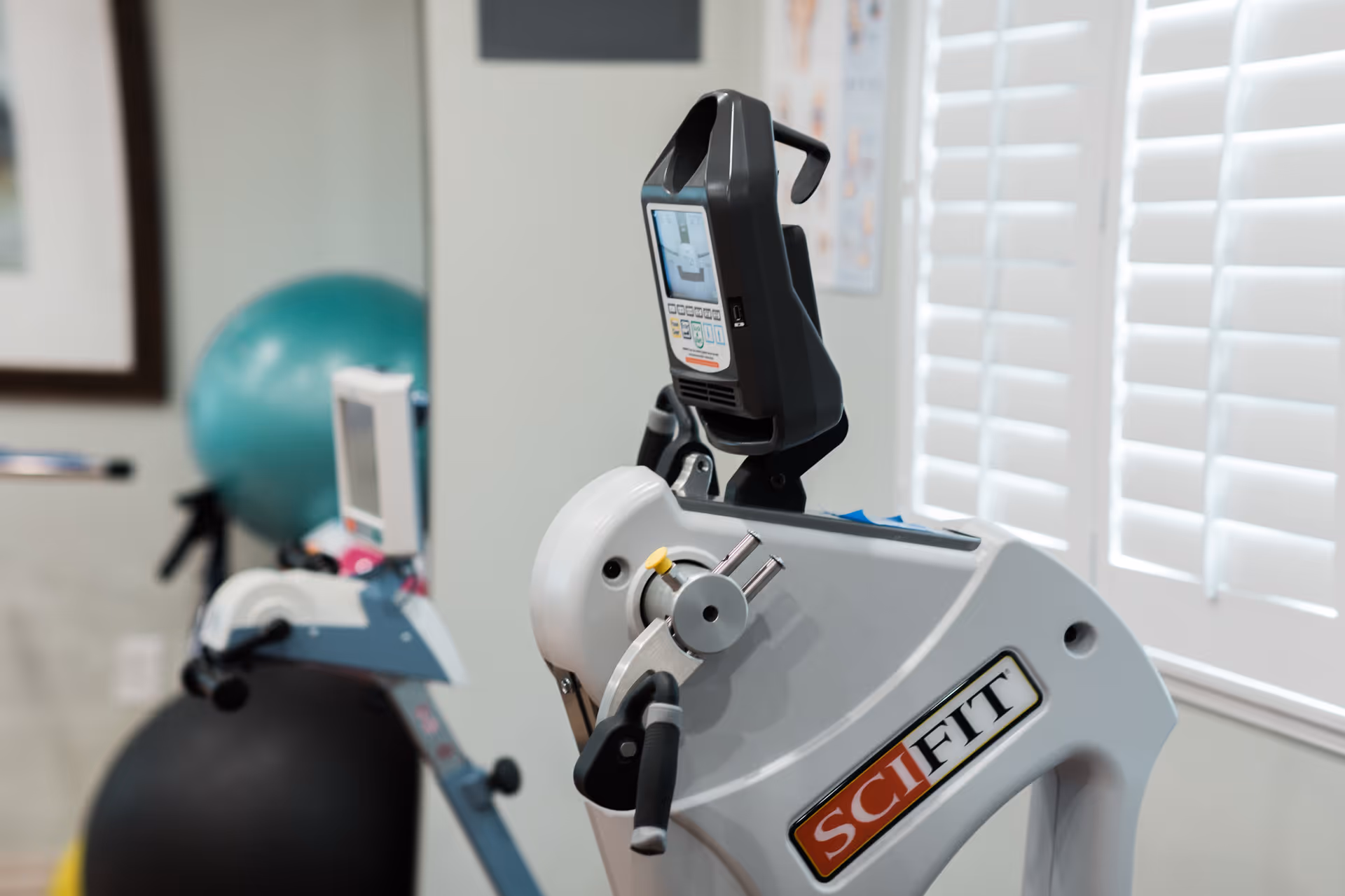 Close-up view of a SciFit exercise machine in a fitness or rehabilitation room with a large exercise ball and another piece of exercise equipment in the background near a window with white blinds.