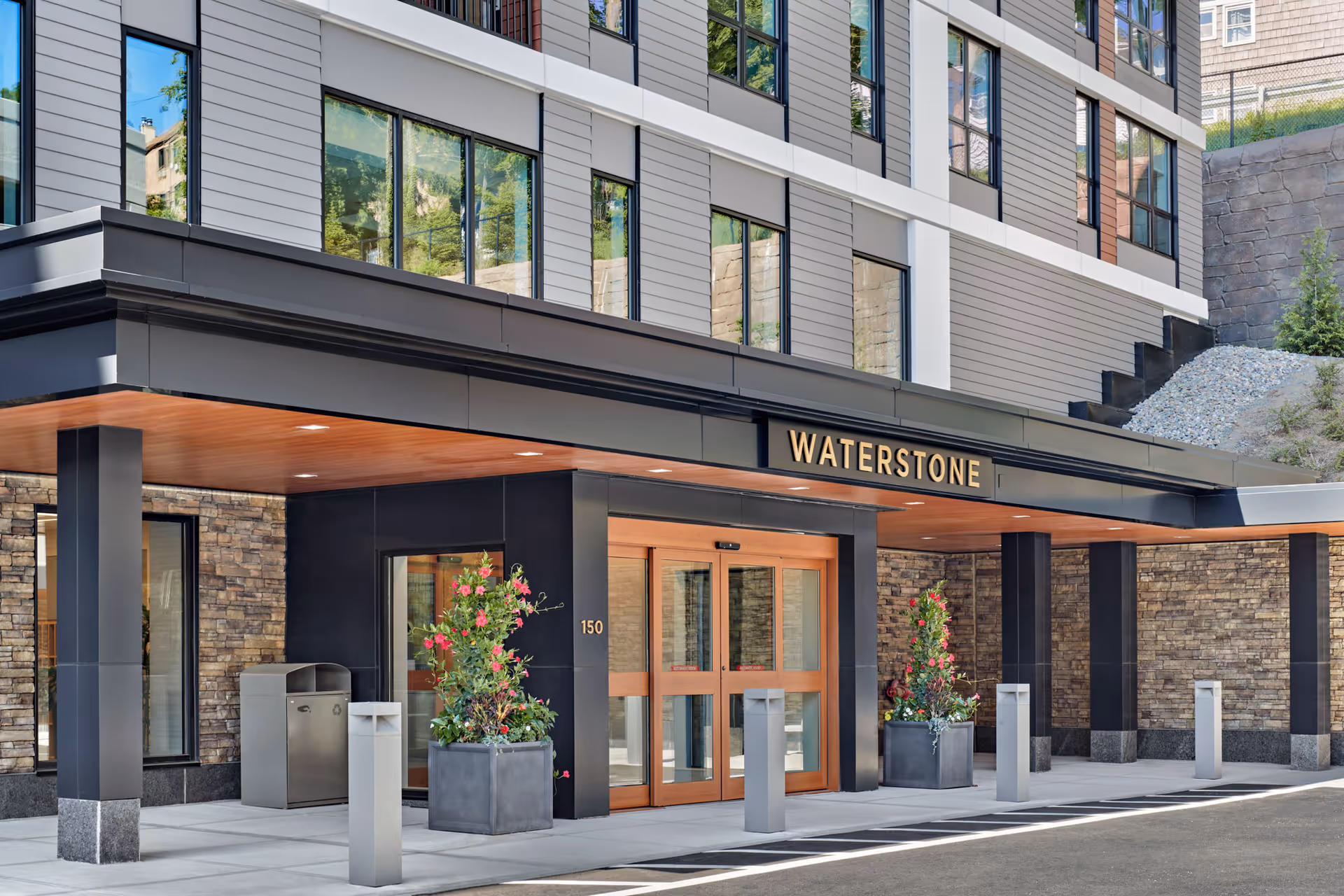 Entrance of the Waterstone senior living facility with a modern exterior featuring large windows, stone walls, black pillars, and a covered drop-off area with automatic sliding glass doors and decorative planters with flowers.