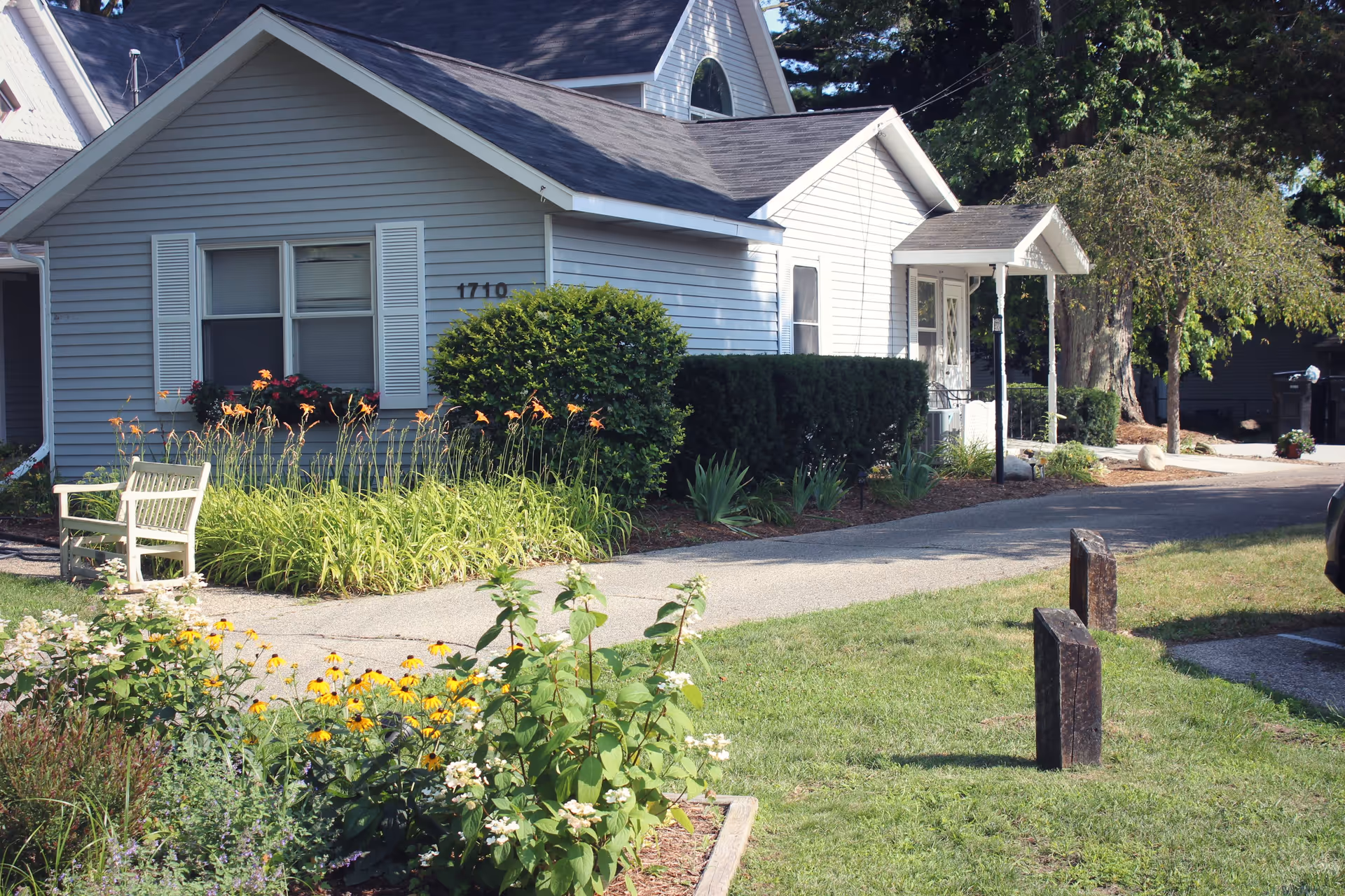 Front exterior of a single-story light-gray house with a small covered porch, flower beds, a bench, and a driveway.