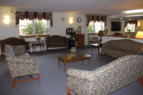 A spacious seating area with patterned sofas and armchairs arranged around coffee tables in a senior living facility common room.