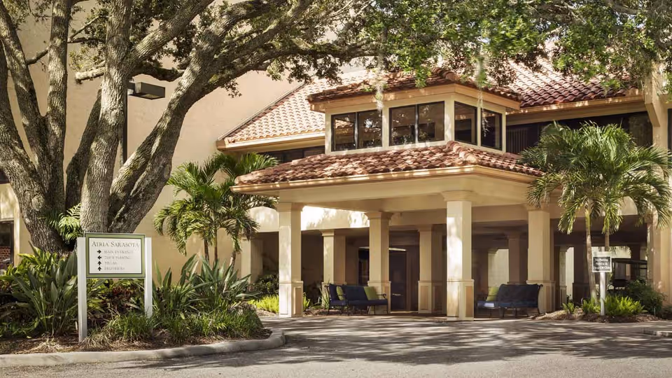 Exterior view of a senior living facility entrance with a covered drop-off area supported by columns, surrounded by palm trees and landscaping. There is a sign indicating directions to the main entrance, visitor parking, and elevators.