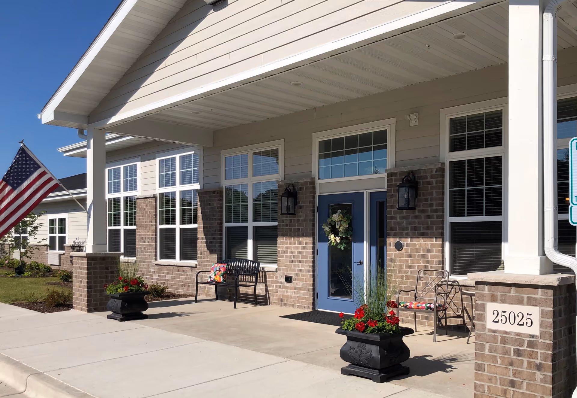 Front entrance of a senior living building with a covered porch, blue door adorned with a wreath, benches, potted flowers, and an American flag.