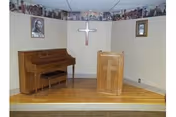 Small chapel area with an upright piano and wooden lectern on a raised platform beneath a wall-mounted cross and framed religious pictures.