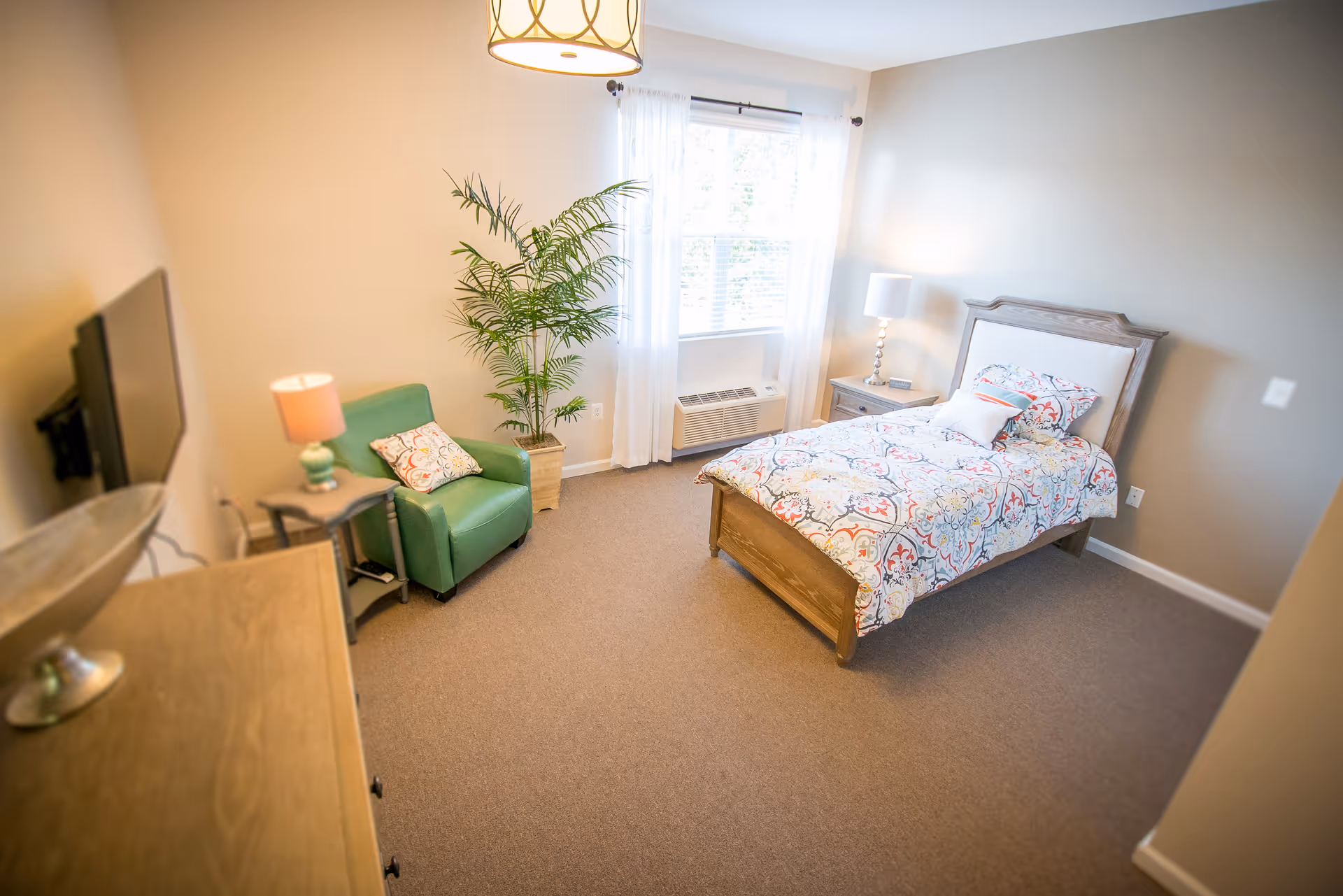 A bright furnished bedroom with a single bed with patterned bedding, a green armchair, potted plant, bedside tables and a wall-mounted TV.