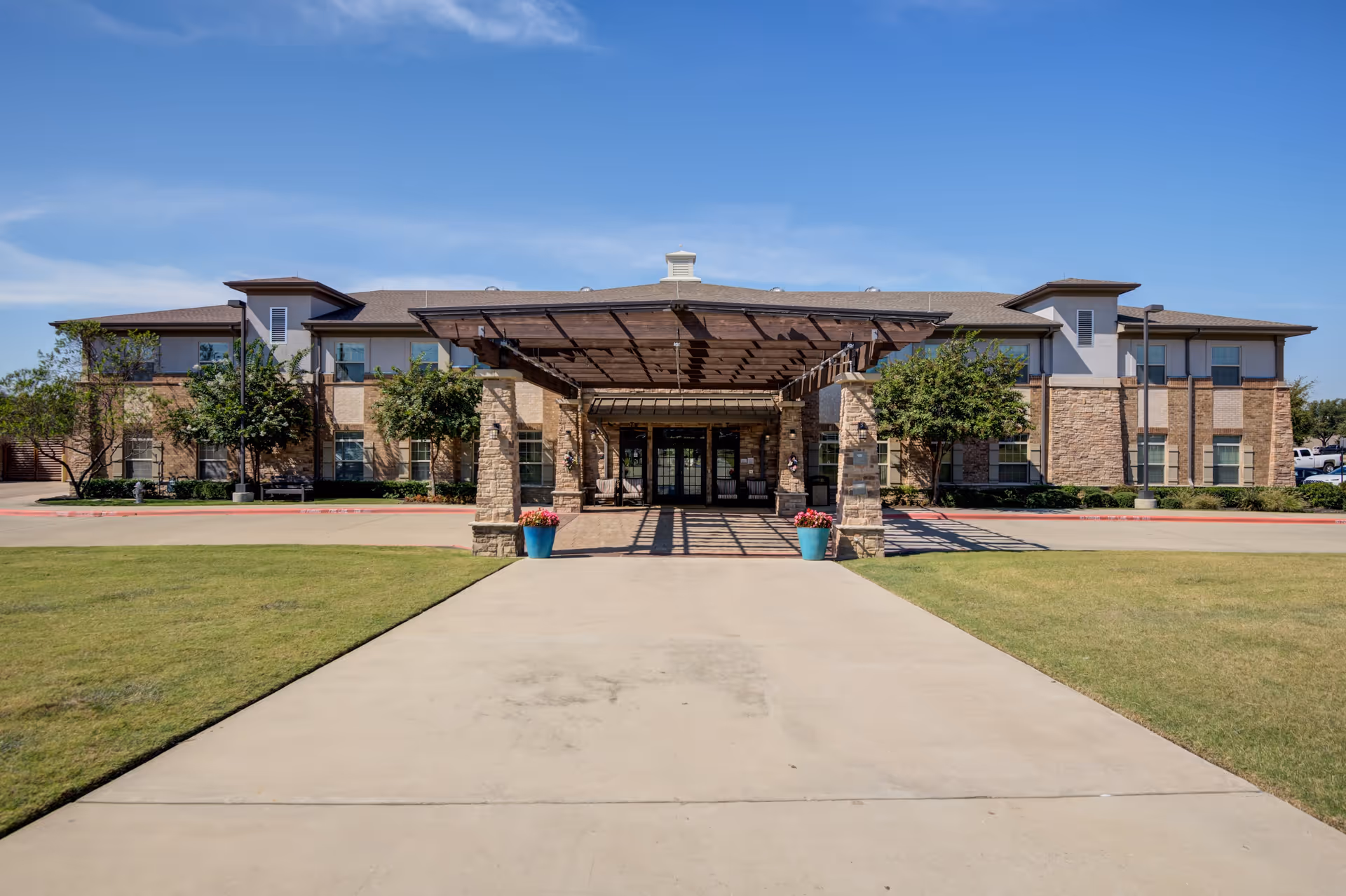 Front exterior view of Truewood by Merrill, Keller, a two-story building with a covered entrance supported by stone pillars, surrounded by green lawns and trees under a clear blue sky.
