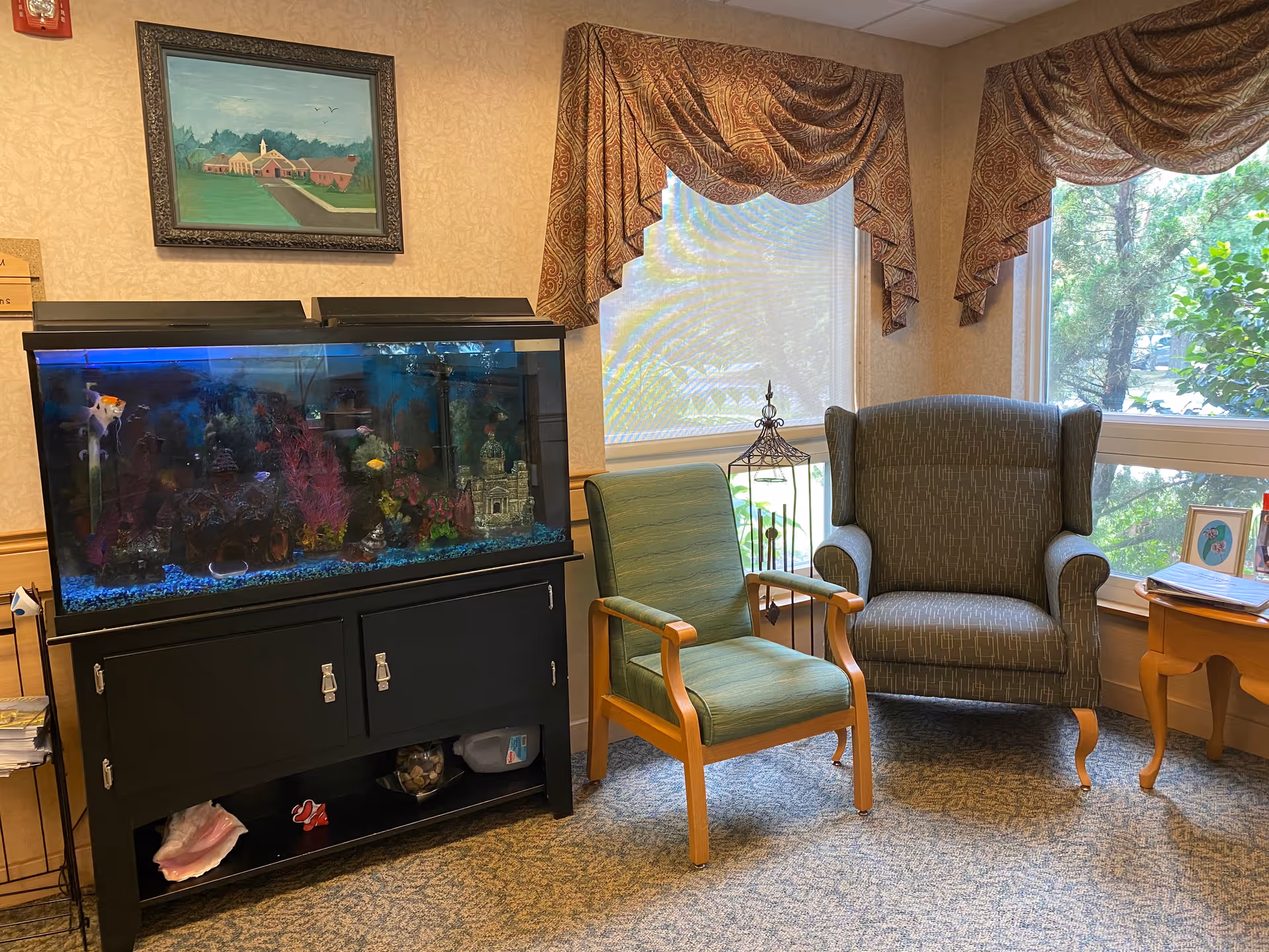 A cozy sitting area with a large aquarium, two upholstered chairs, a side table, and windows with draped curtains.