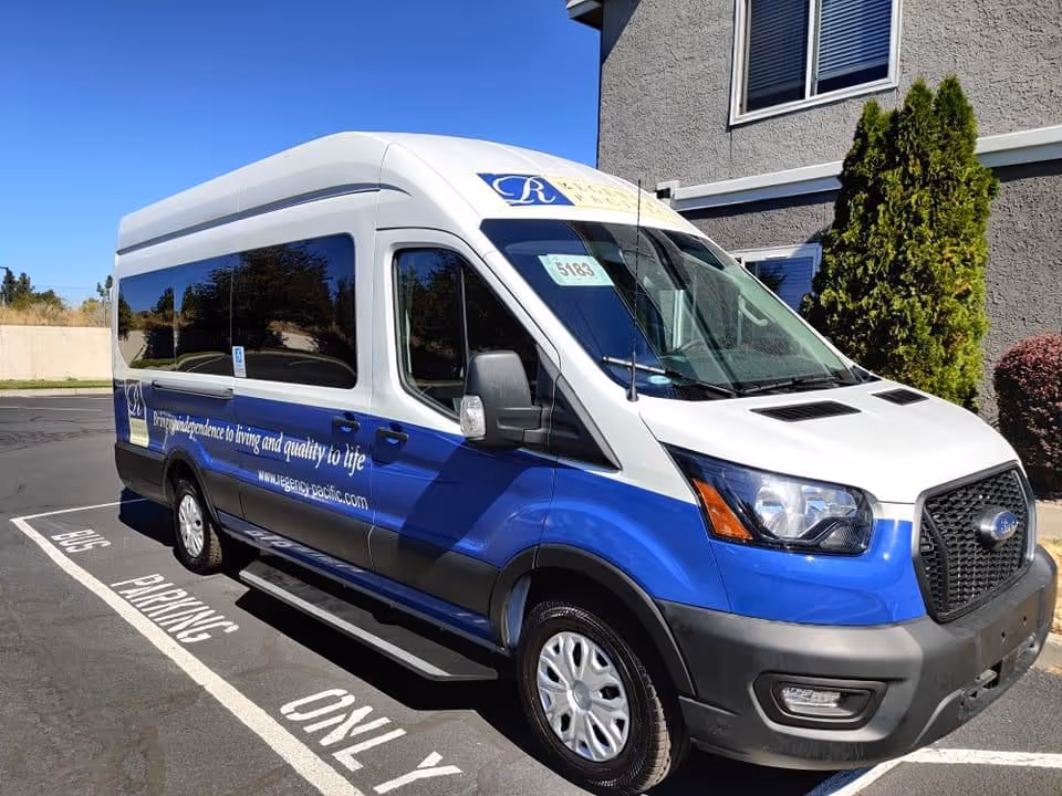 A white and blue shuttle van parked in a designated bus parking spot outside a building, with clear skies and some greenery nearby. The van has signage indicating it is associated with Regency Village at Bend.
