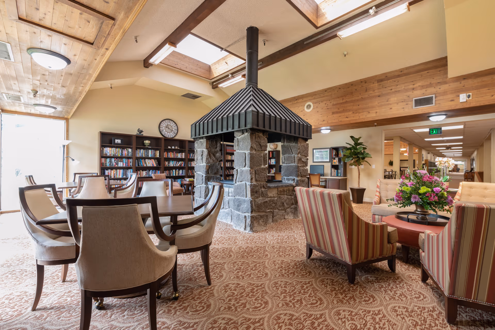 A cozy common area in a retirement community featuring a central stone fireplace with a black metal chimney, surrounded by comfortable upholstered chairs and a round table with a floral arrangement. There are bookshelves filled with books against the wall, a clock, and a skylight letting in natural light. The room has a patterned carpet and wooden ceiling accents.